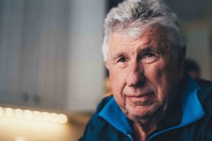 White-haired man wearing a blue top looking into the distance