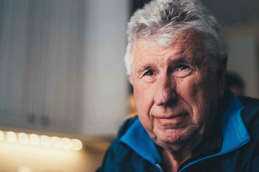 White-haired white man wearing a blue top looking into the distance