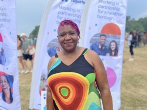 Picture of Annmarie smiling, outside the Lambeth Together tent at the Lambeth Country Show. She's wearing a swirly bright black, green and orange top and has short pink braids