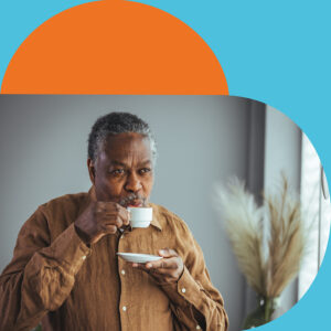 An older mixed race gentleman holds a tea cup and saucer and blows his drink to cool it down