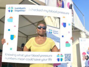 Yvette outside the Lambeth Together tent at the Lambeth Country Show. She's holding a frame which says 'I've checked my blood pressure' and wears a yellow top and sunglasses