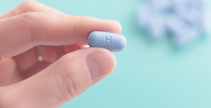 A close-up of a hand holding a small, oval-shaped blue PrEP pill with the letter "H" imprinted on it, used for HIV prevention. 