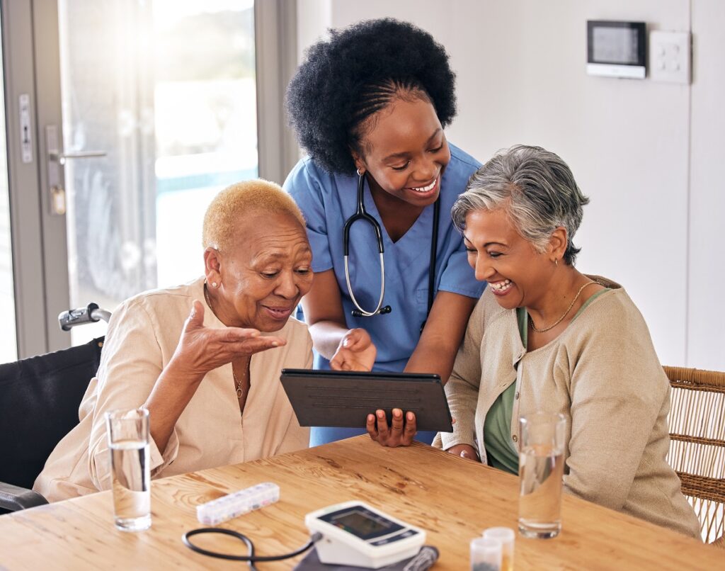 A nurse in the kitchen of a retirement home, showing information on a tablet to two older female friends