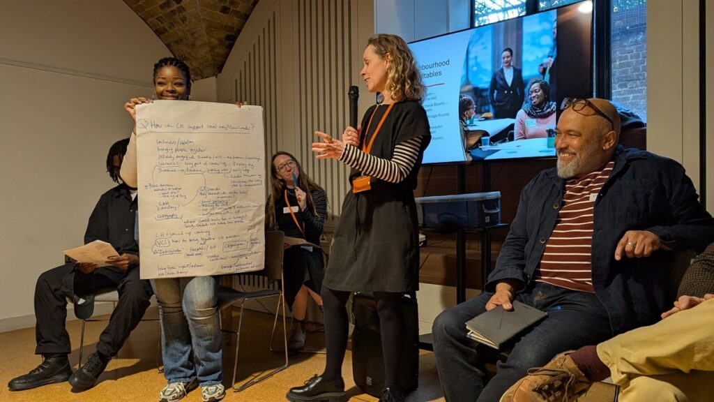 Attendee at the Creative Health in Lambeth’s Neighbourhoods event speaking while another holds a large sheet of handwritten notes; others seated with notebooks and a presentation screen in the background.