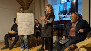Attendee at the Creative Health in Lambeth’s Neighbourhoods event speaking while another holds a large sheet of handwritten notes; others seated with notebooks and a presentation screen in the background.