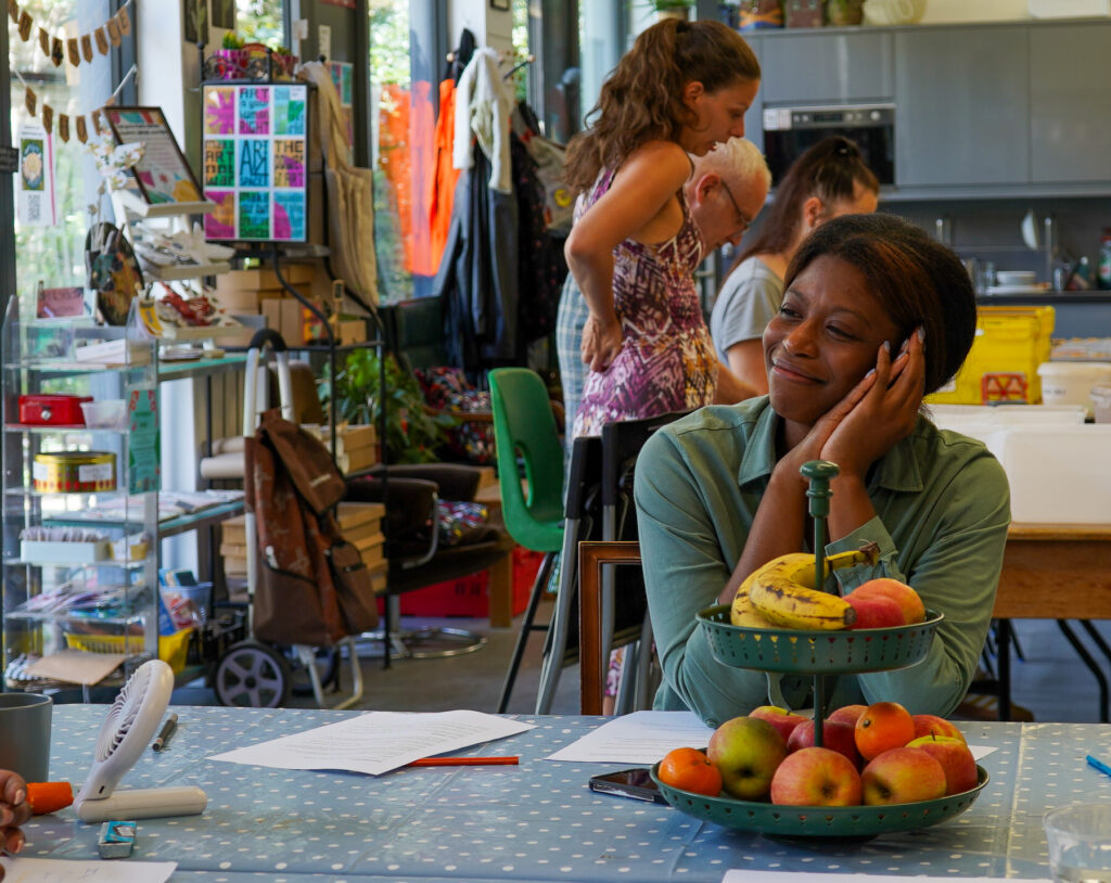 A lady sits at a table with fruit and papers in the foreground, while two people stand and talk in a creative studio space.