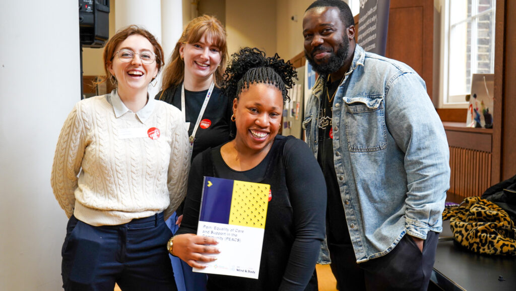 Four people standing indoors, one holding a colorful booklet with text, near a table and window.