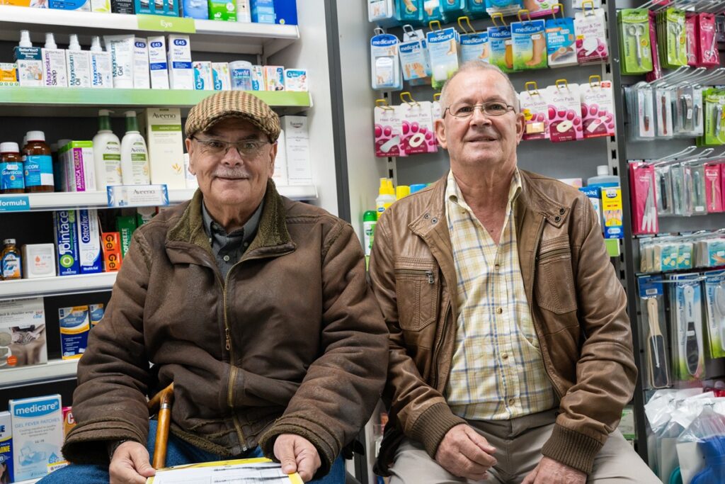 Two friends, Jose and Joquim, sit in their local pharmacy, with medicine shelves behind.
