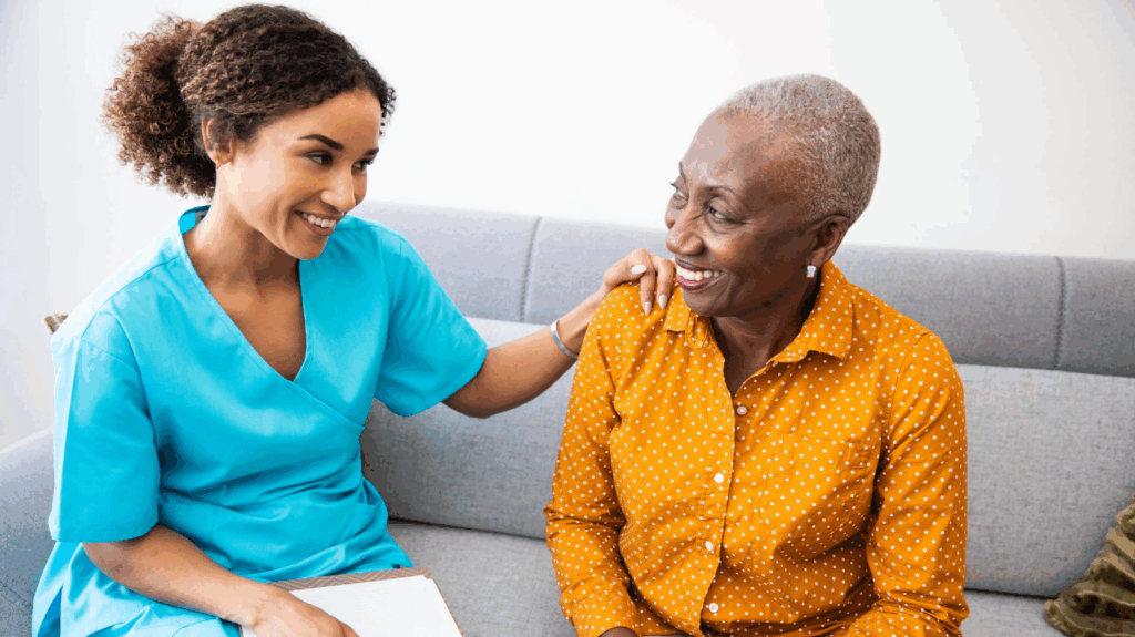 Female healthcare worker sitting beside an older woman on a sofa, offering support with a hand on her shoulder.