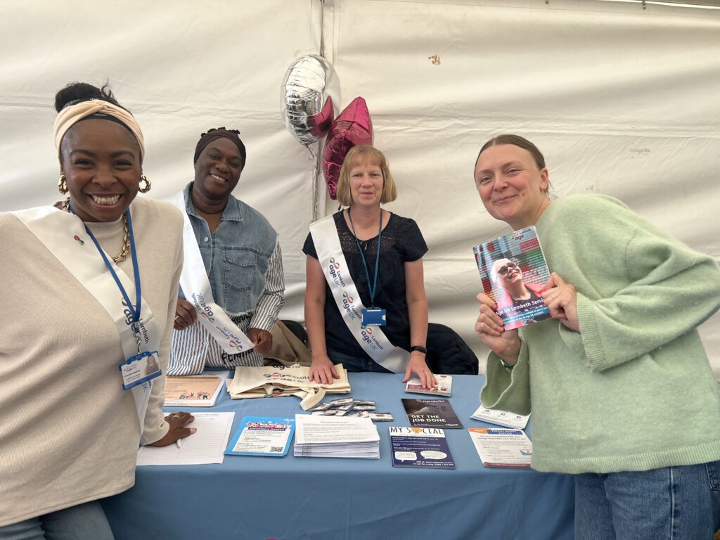 Four smiling Age UK Lambeth advisers at their information stall in a marquee at a community event