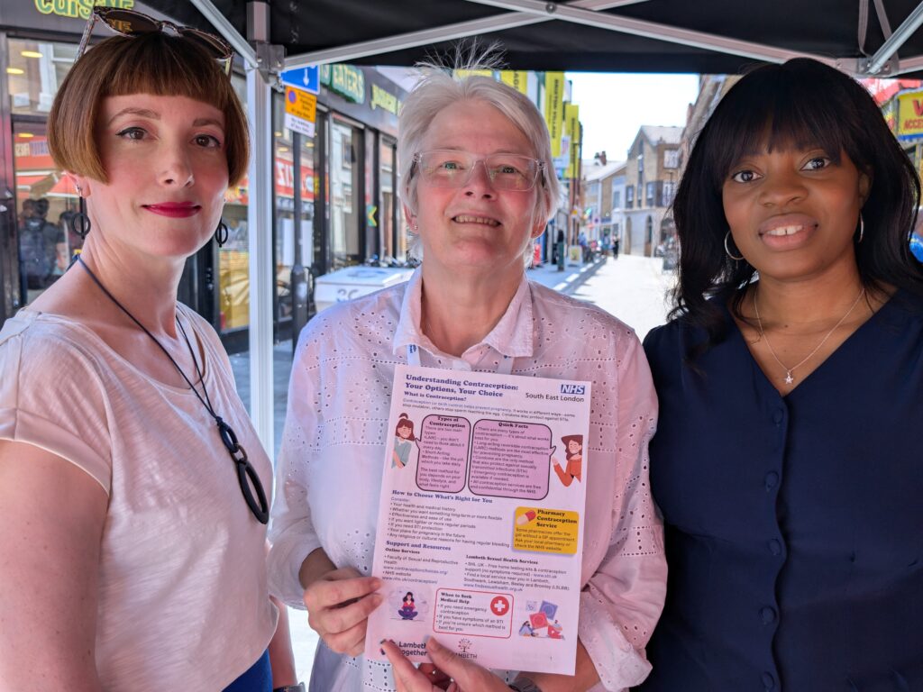 Three women standing under a gazebo on a busy street, holding a leaflet about women's health information.