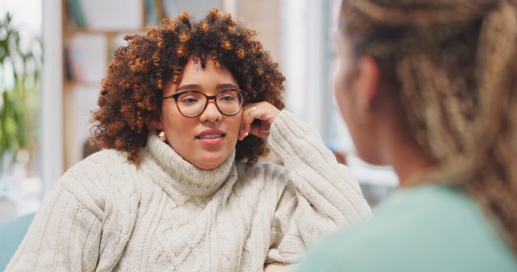 Two women sit indoors facing each other in conversation, one in a light cable-knit sweater and the other with braided hair, with bookshelves in the background.