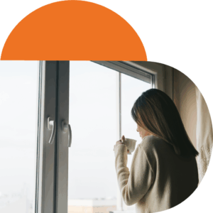 A woman standing by a large window holding a mug, wearing a light sweater, with soft daylight coming through