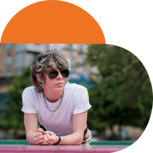 Non-binary person with short wavy hair wearing a white t-shirt and chain necklace, leaning forward on a colorful surface outdoors with trees and buildings in the background