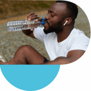 Person wearing a white t-shirt sitting outdoors on a gravel path, holding a clear plastic water bottle and drinking, with wireless earbuds in