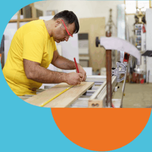 Person in a yellow shirt measuring a wooden plank with a tape measure and marking it with a pencil in a workshop.