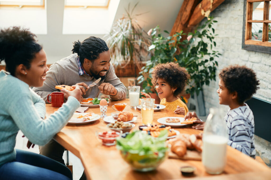 Happy black parents and their kids talking while having breakfast together in dining room.