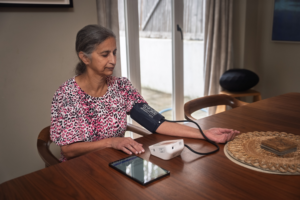 An older woman sits at a table checking her blood pressure with an electronic monitor. She is wearing a pink patterned top. A tablet lies on the table beside her. She appears calm and focused.