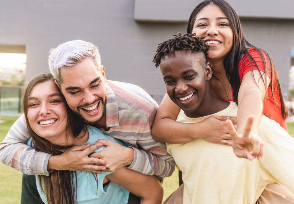 Four friends are outside, two giving piggyback rides, laughing and making a peace sign. 
