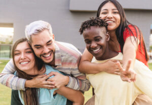 Four friends are outside, two giving piggyback rides, laughing and making a peace sign.