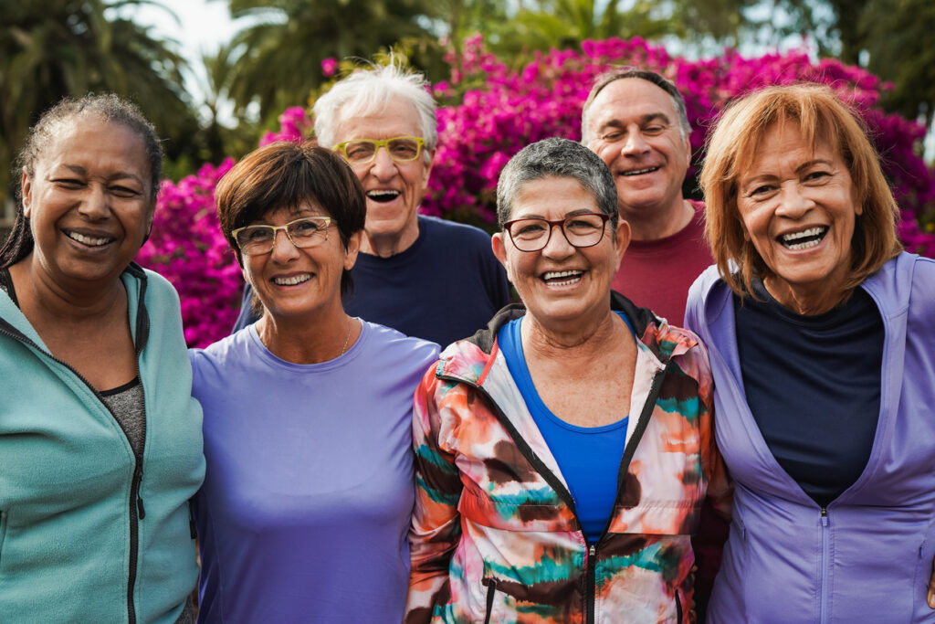 A group of six older adults stand closely together outdoors, smiling and laughing, with vibrant pink flowers and green foliage in the background.