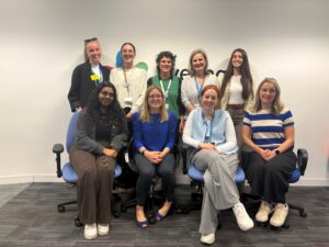 A group of ten women, some seated and some standing, pose and smile for a photo in an office setting with a plain white wall and two blue chairs.