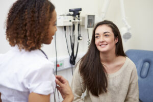 A young woman sits in a medical examination room, smiling and talking with a healthcare professional in a white coat. Medical equipment is visible on the wall behind them.