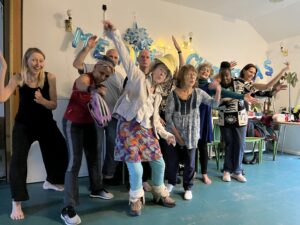 A group of people of various ages, including members from Unbound Moving Arts and Age UK Lambeth, joyfully dance and pose in a festive indoor setting with blue “Merry Xmas” decorations and fairy lights on the wall behind them.