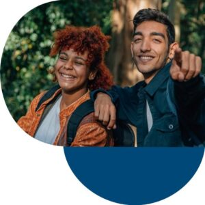 Two young people with rucksacks smiling outdoors; one has curly ginger hair and an orange jacket, the other has short dark hair and points towards the camera, both appear happy and relaxed with greenery in the background.