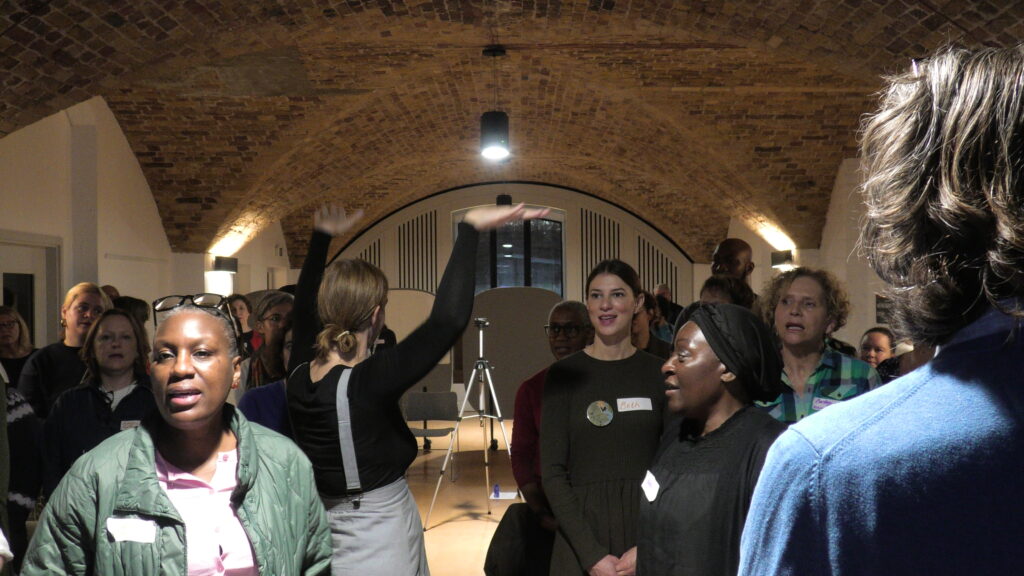 A group of people stand together in an arched brick room, some talking and some singing. One person in the centre raises an arm as others face different directions, creating a lively atmosphere.