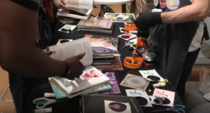 Two people look through books and art supplies on a cluttered table during Mental Health Awareness Week Lambeth 2026. Books, zines, circular paper cut-outs, scissors, and a badge-making machine cover the surface; only their hands and arms are visible.