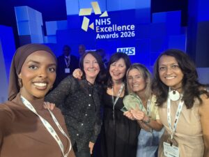 Five women from Lambeth Women’s and Girls’ Health Hub smile and pose for a selfie at the NHS Excellence Awards 2026, standing in front of a blue-lit stage with the NHS logo and awards signage in the background.