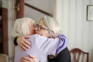 Two elderly women with grey hair warmly hugging each other indoors. One woman wears glasses and a dark jumper, while the other wears a light purple top. They both appear happy and content in the embrace.