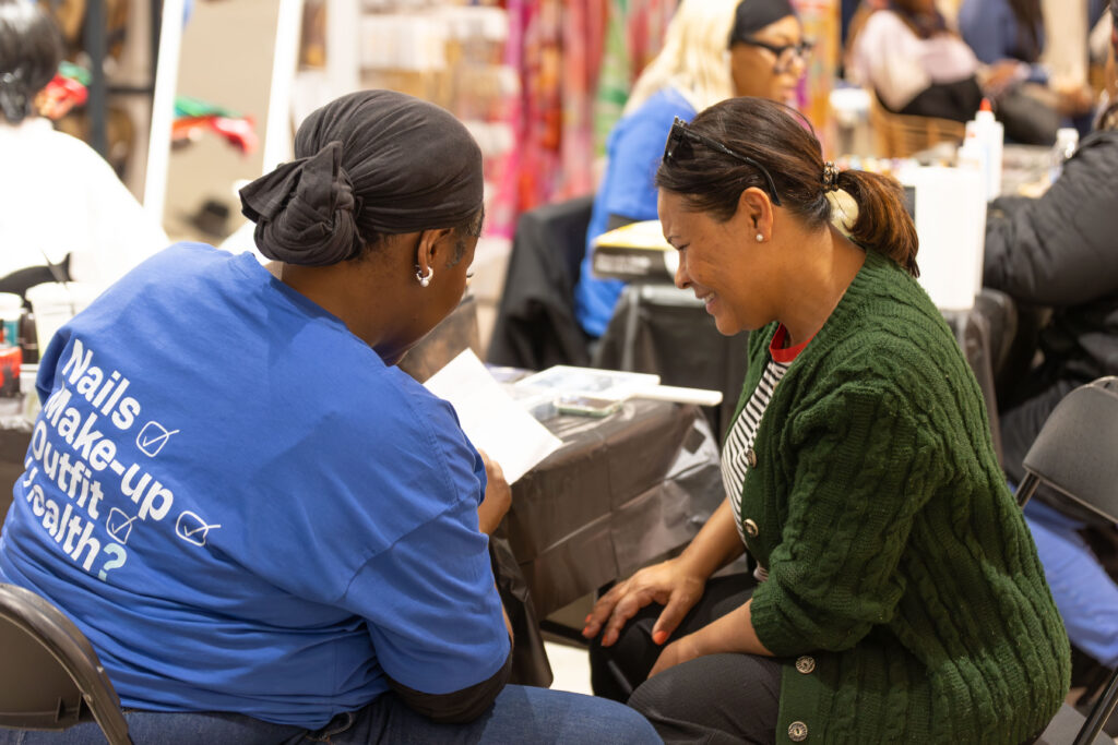 Two women sit opposite each other at a table, one wearing a blue shirt reading Nails, Makeup, Outfit, Health?, as they look at a piece of paper and smile in a bustling indoor setting.