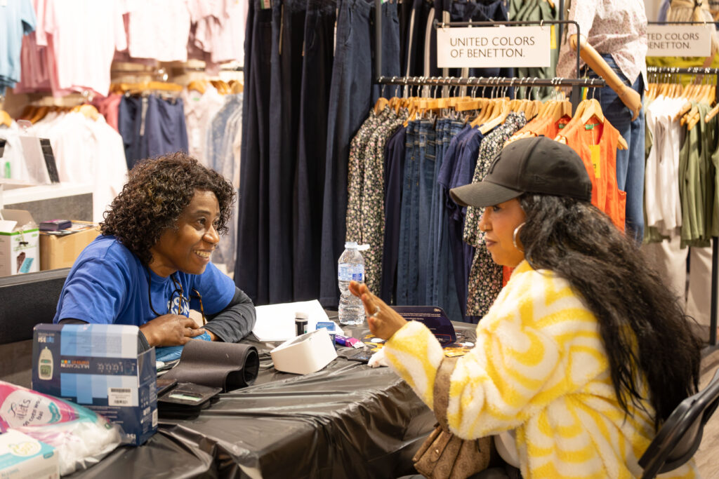 Two women sit across a table in a clothes shop, having a friendly conversation. One wears a blue shirt and smiles, while the other wears a yellow patterned jacket and cap, gesturing with her hand. Clothes hang in the background.