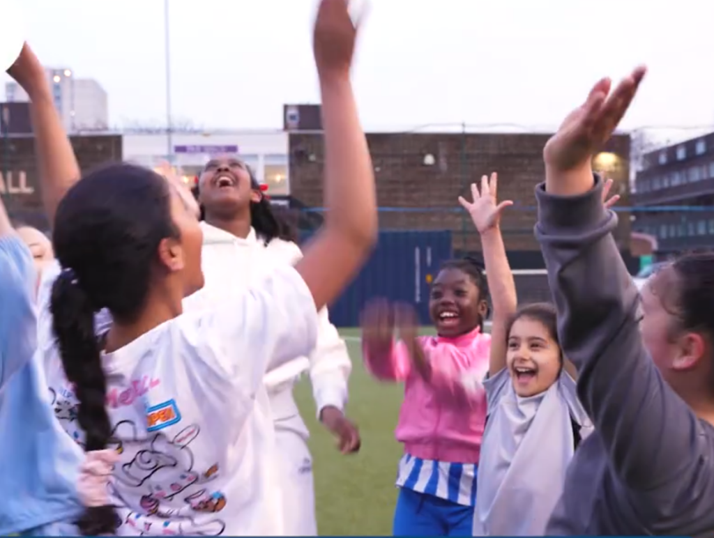 A diverse group of girls celebrate together on an outdoor sports pitch, raising their arms and smiling. Logos for Lambeth Together and Neighbourhood and Wellbeing Alliance are visible. Text reads: “Women’s and Girls’ Health Hub, Lambeth.”.