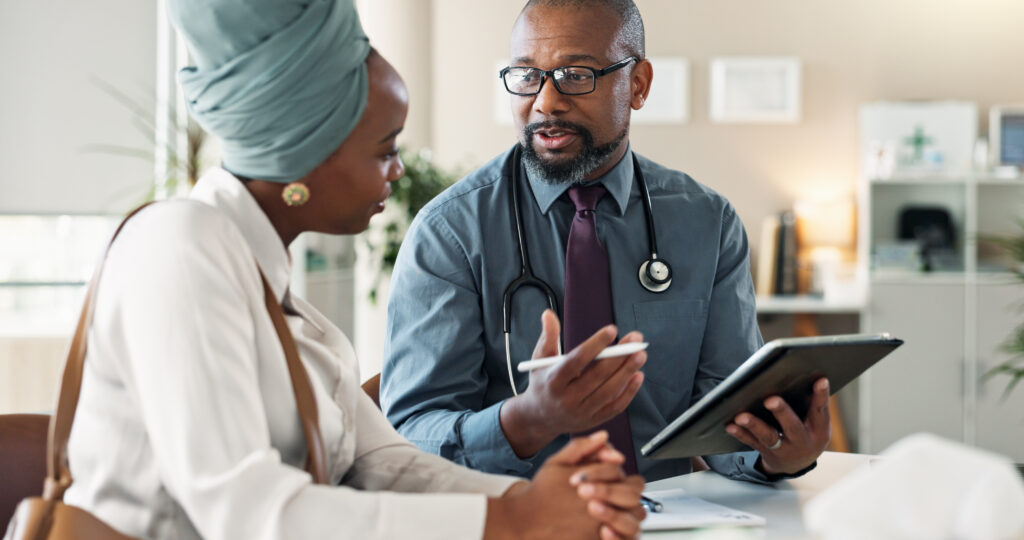 A doctor wearing a stethoscope speaks with a patient in a headscarf whilst holding a digital tablet. They are seated at a desk in a bright, modern office.