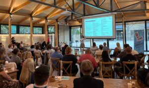 A speaker presents to a seated audience in a modern venue with wooden beams and large windows. A large screen displays a slide, and attendees are listening attentively.