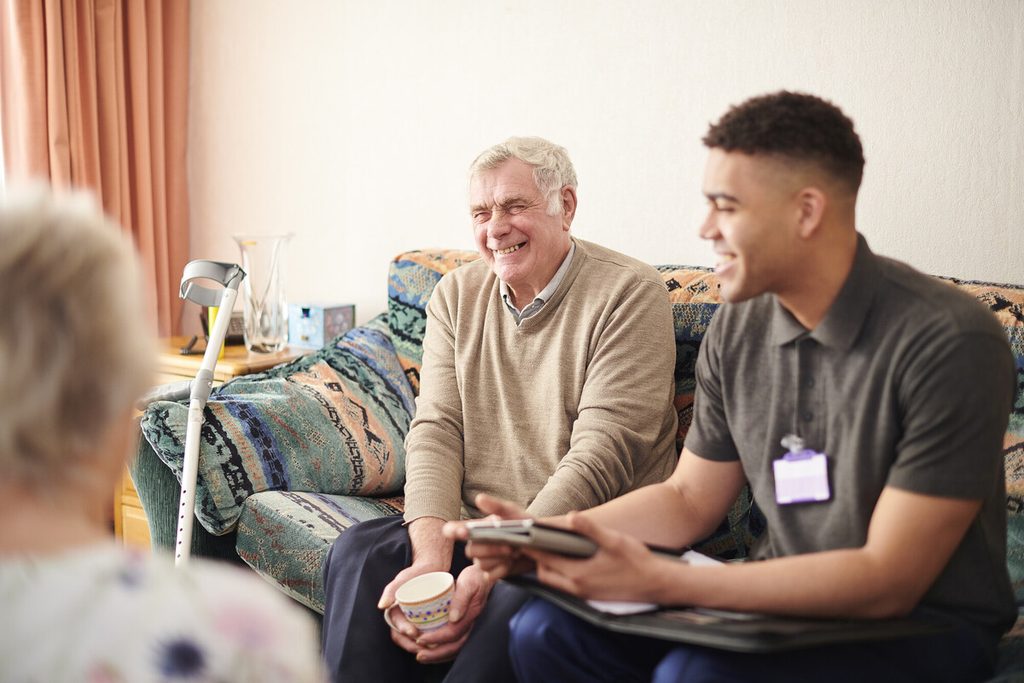 A smiling elderly man sits on a sofa with a carer who is holding a tablet.